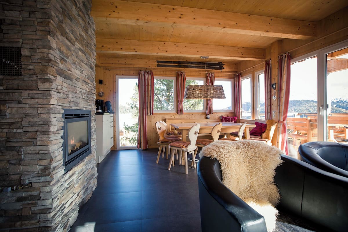 A cozy room with a stone fireplace, wooden ceiling, and large windows offering a view of the outdoors.