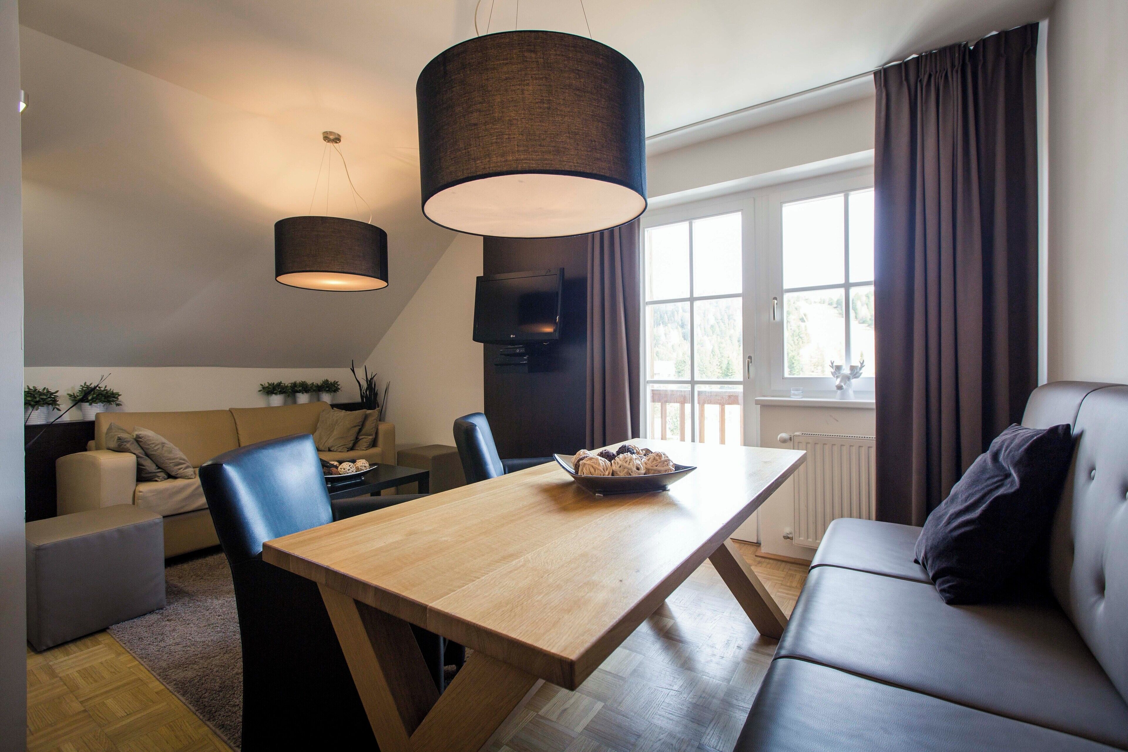Un salón moderno con una mesa de comedor de madera, sillas negras, un sofá y un amplio ventanal con cortinas.