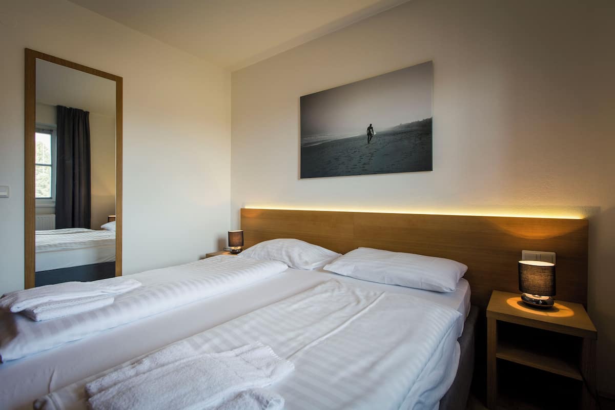 A hotel room with two beds, a large mirror, a nightstand with a lamp, and a painting of a person on a beach.