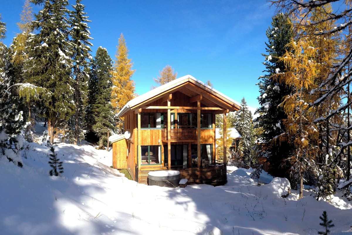 A wooden cabin with a balcony, surrounded by snow-covered trees and a clear sky.