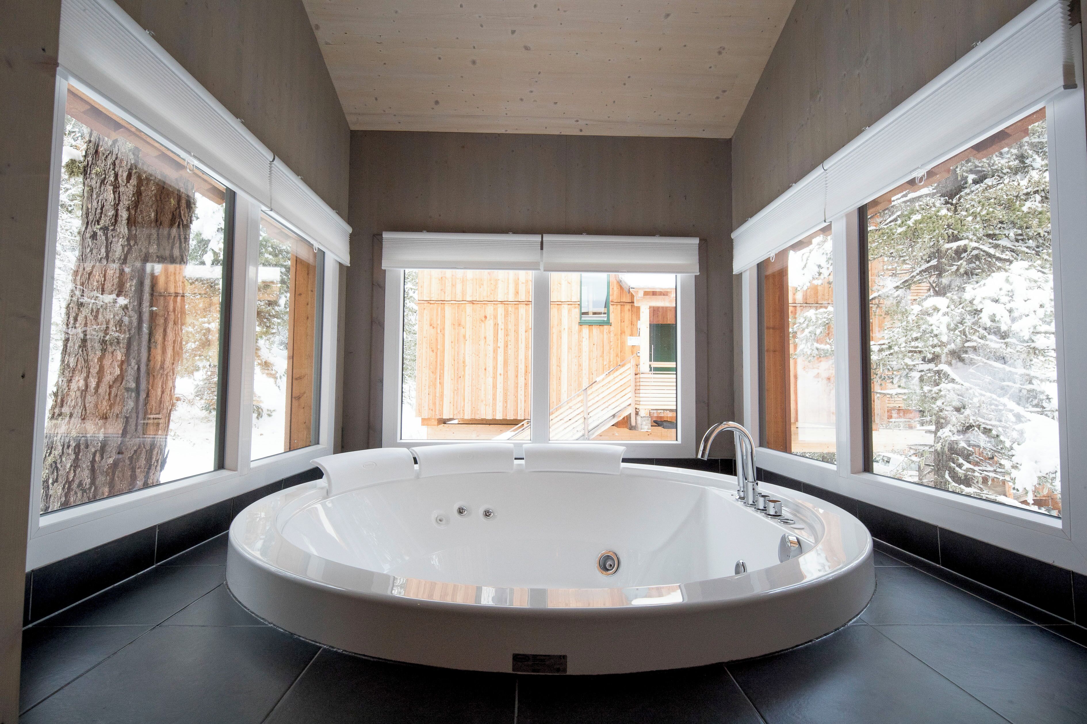 Un baño moderno con una bañera grande, ventanas amplias y vistas a una terraza de madera y árboles.