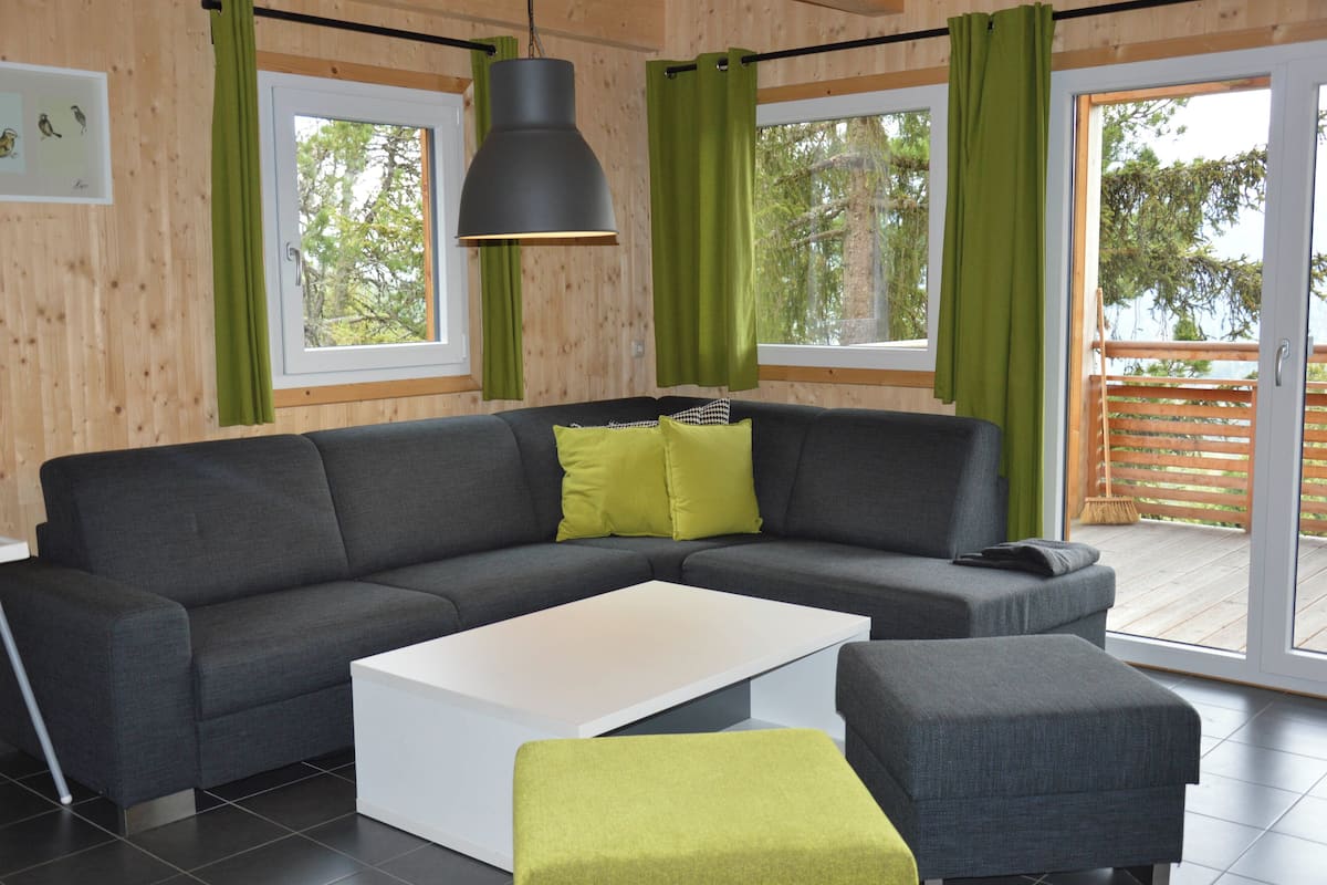 A modern living room with a grey sectional sofa, a white coffee table, and a green ottoman. There are two windows with green curtains, a black pendant light, and a wooden deck visible through the glass doors.