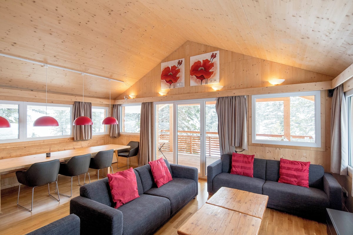 A modern cabin interior with wooden ceilings, large windows, a dining area, and seating arrangements.