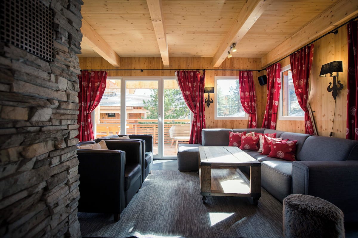 A cozy room with a stone wall, wooden ceiling, a sofa with red cushions, a coffee table, and a view through large windows.