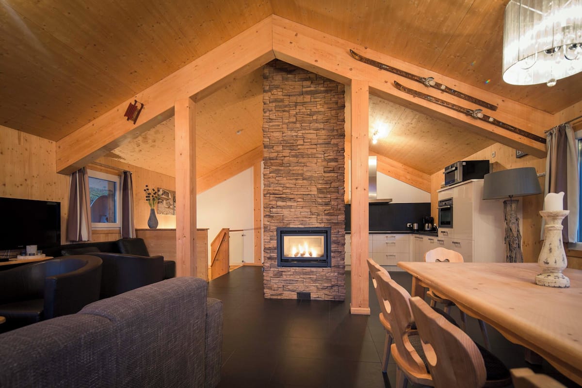 A cozy living room with a stone fireplace, wooden beams, and a dining area with a table and chairs.