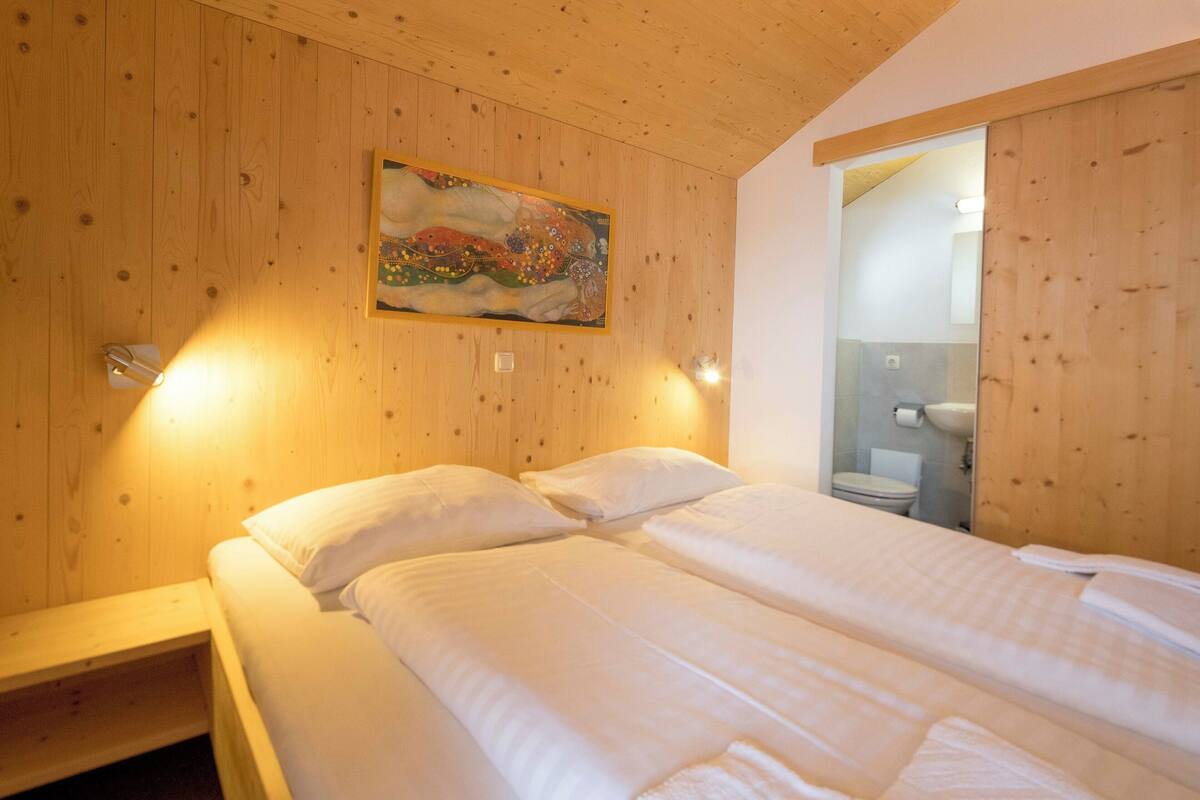A wooden-paneled room with a bed, a painting, and a bathroom visible through an open door.