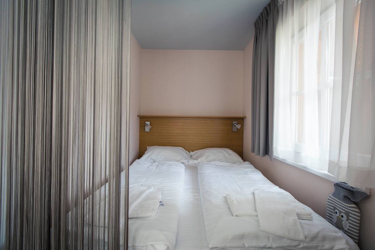 A double bed with white linens, a wooden headboard, and a window with sheer curtains.