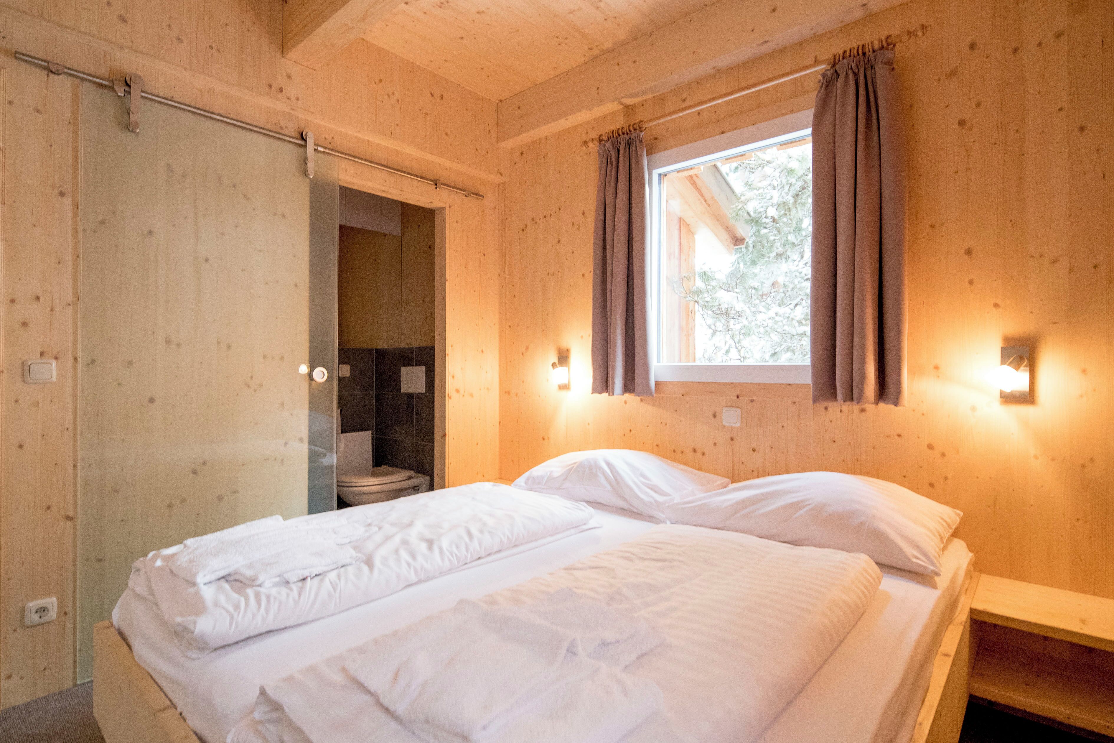 A wooden cabin bedroom with a bed, a window with curtains, and a bathroom visible through an open door.