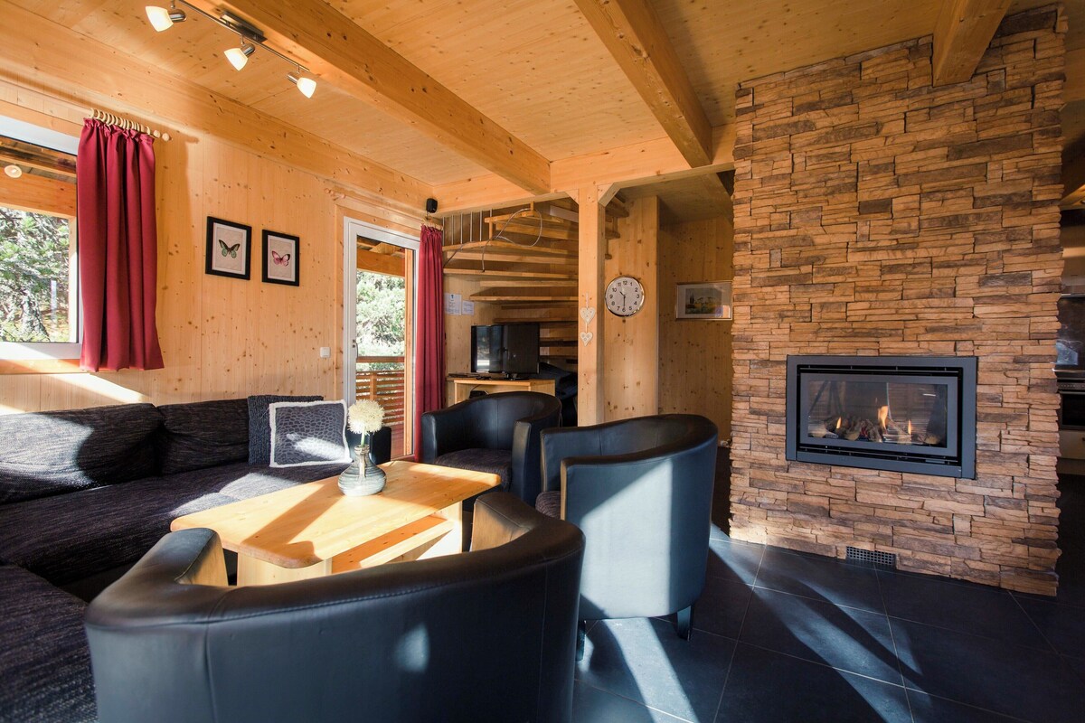 A cozy cabin interior with a stone fireplace, wooden ceiling beams, a sofa, and a dining table.