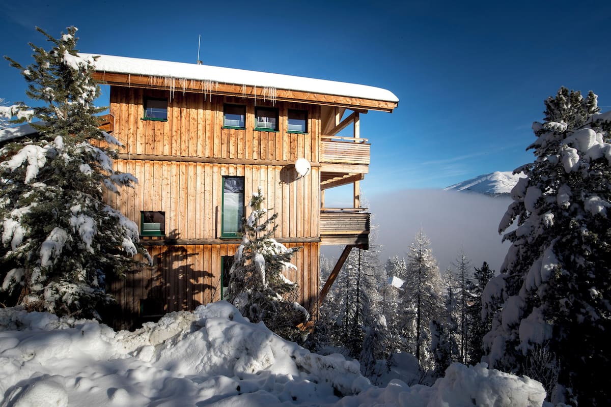 A wooden cabin with multiple levels, surrounded by snow-covered trees and a clear sky.