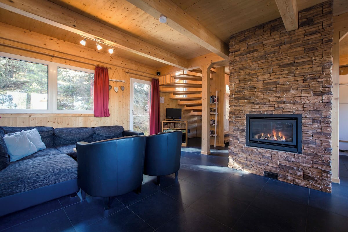 A modern living room with a stone fireplace, wooden ceiling beams, and a large window with curtains.