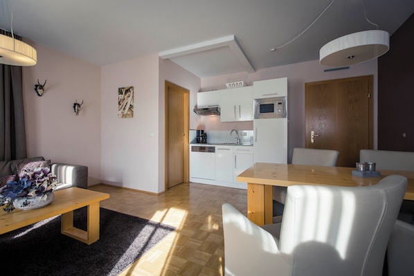 Una sala moderna con un sofá, una mesa de centro y un comedor con mesa y sillas de madera. Se observa una cocina al fondo con armarios blancos y un microondas.