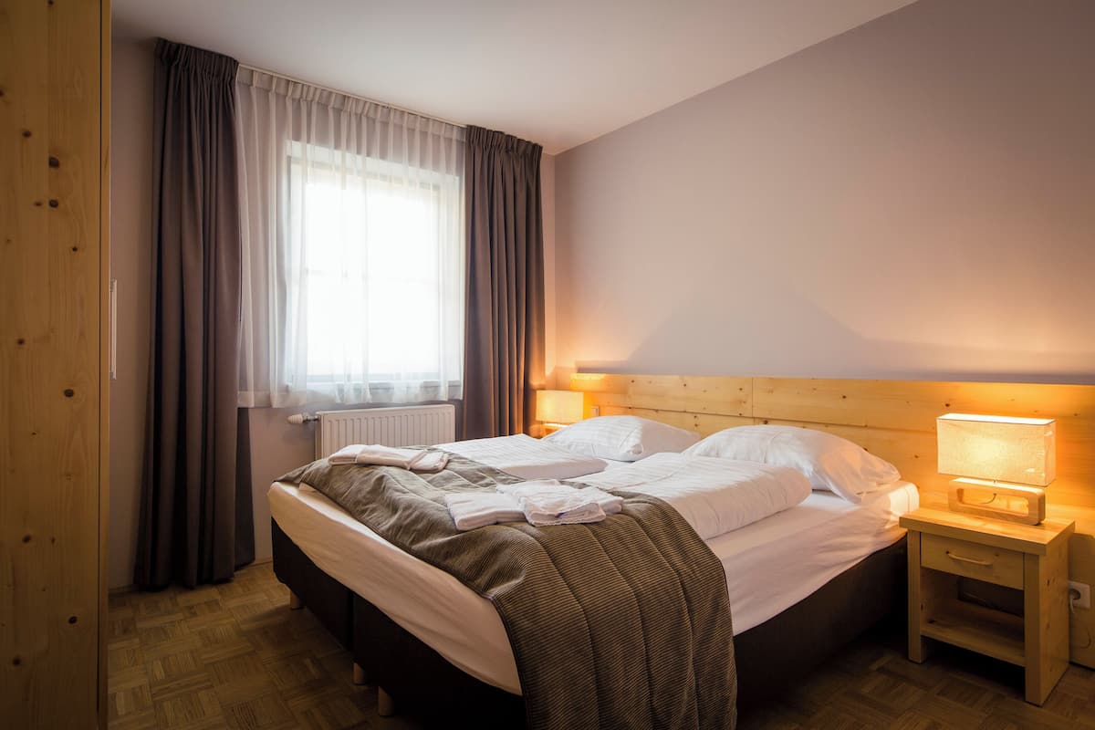 A neatly made bed with a wooden headboard, a bedside table with a lamp, and a window with curtains.