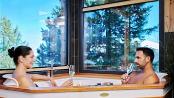 Ein Paar entspannt in einer Whirlpool-Badewanne mit Blick auf Bäume und ein Gebäude im Hintergrund.