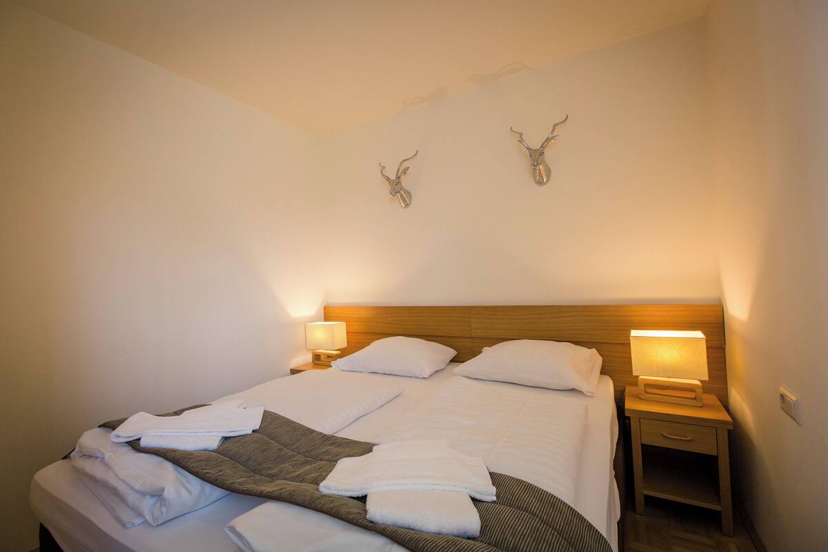 A neatly made bed with pillows and folded towels, two wall-mounted deer antlers, a bedside lamp, and a wooden nightstand with a drawer.