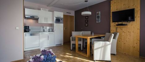 A modern kitchen with white cabinets, a microwave, and a coffee machine. There is a dining area with a wooden table and white chairs. A flat-screen TV is mounted on the wall.