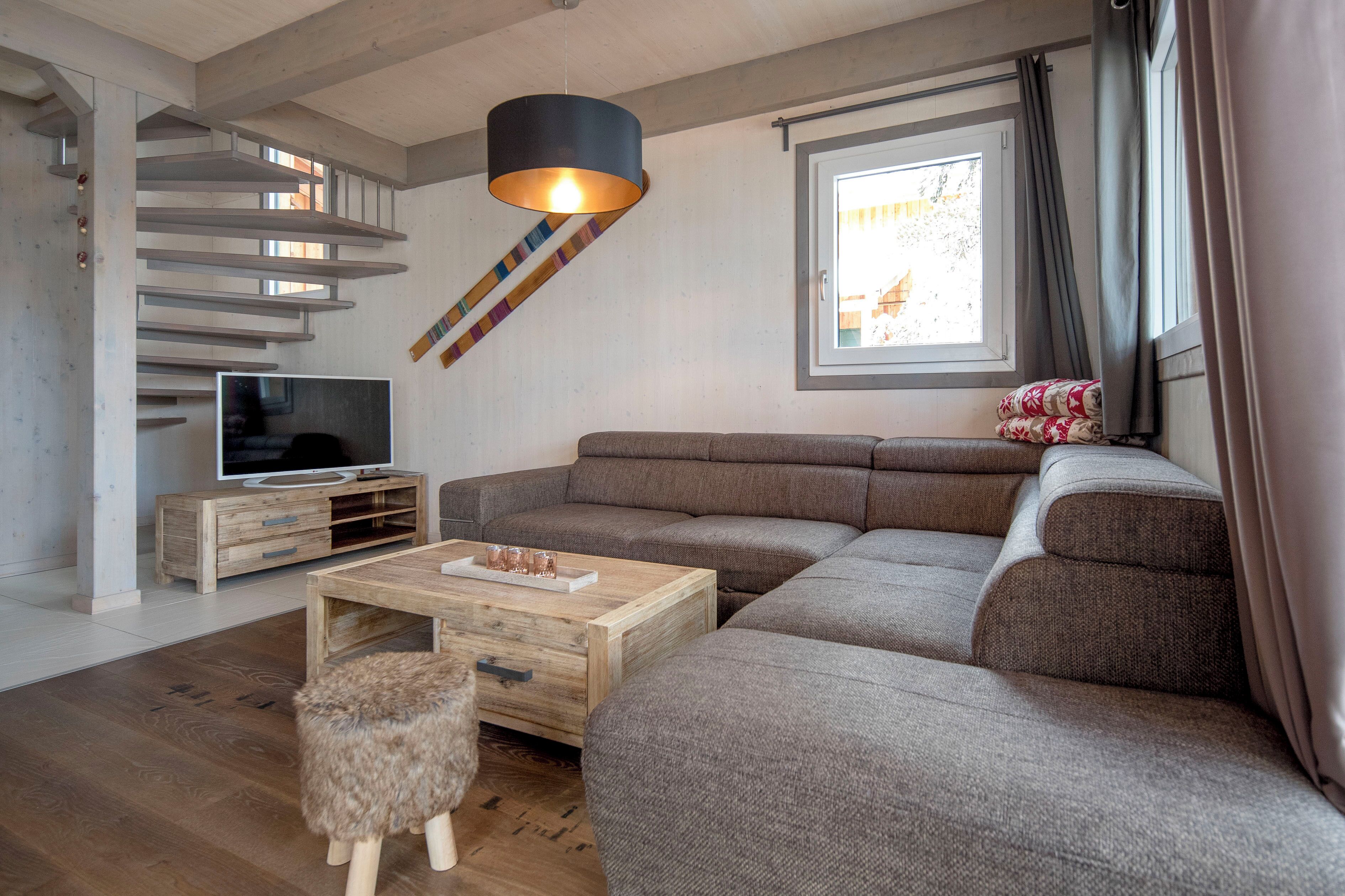 A modern living room with a sectional sofa, a wooden coffee table, a TV on a wooden stand, a staircase with a unique design, and a window with curtains.