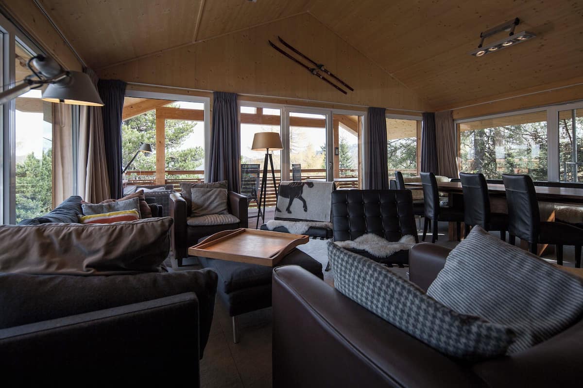 A cozy cabin-style room with a large window, a sofa, a chair, a table, and a ski rack.