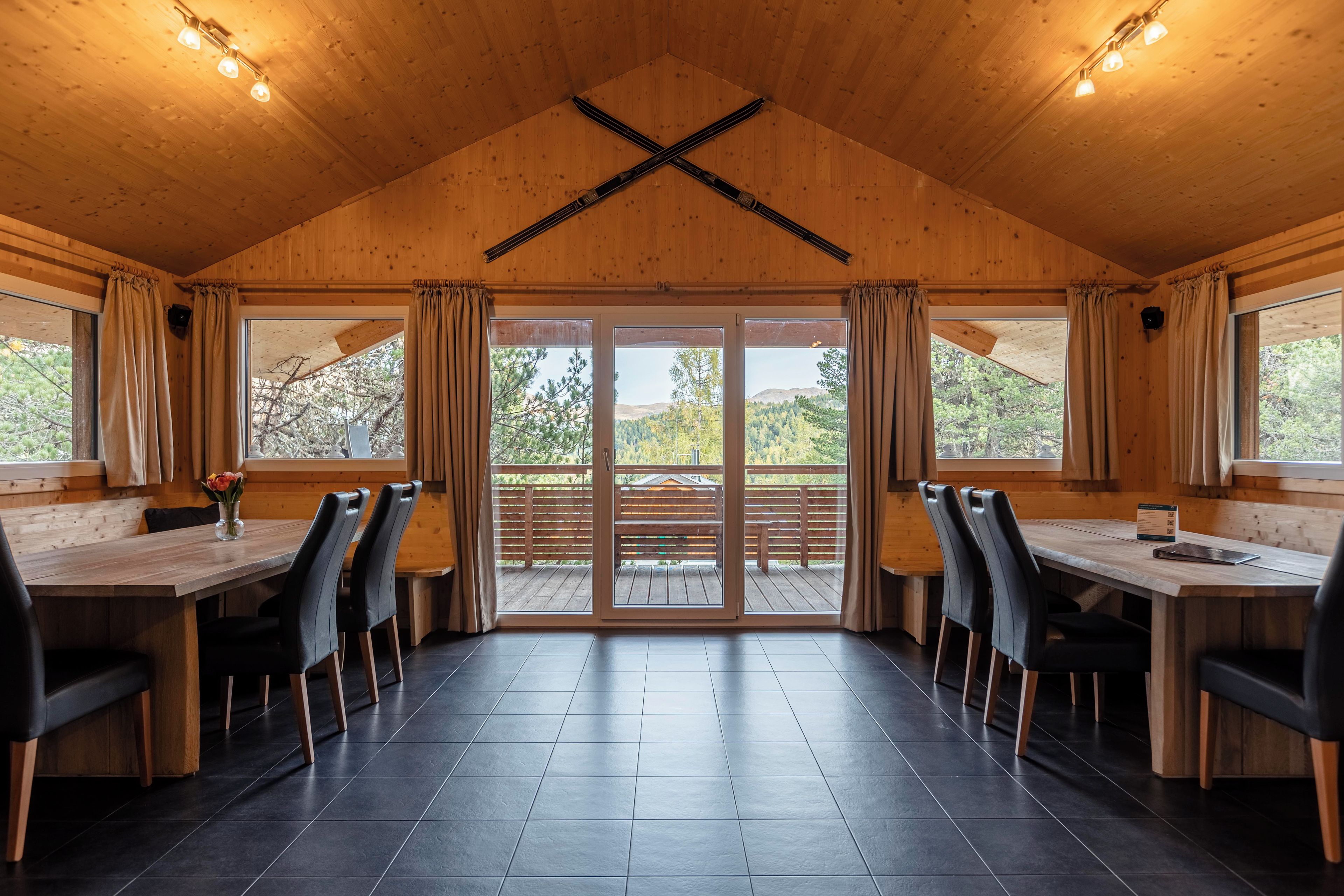 Un interior de madera con un comedor que incluye una mesa larga y sillas, ventanas grandes con cortinas, y vistas a una terraza exterior.