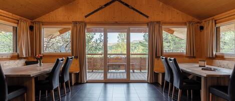 Un interior de madera con un comedor que incluye una mesa larga y sillas, ventanas grandes con cortinas, y vistas a una terraza exterior.