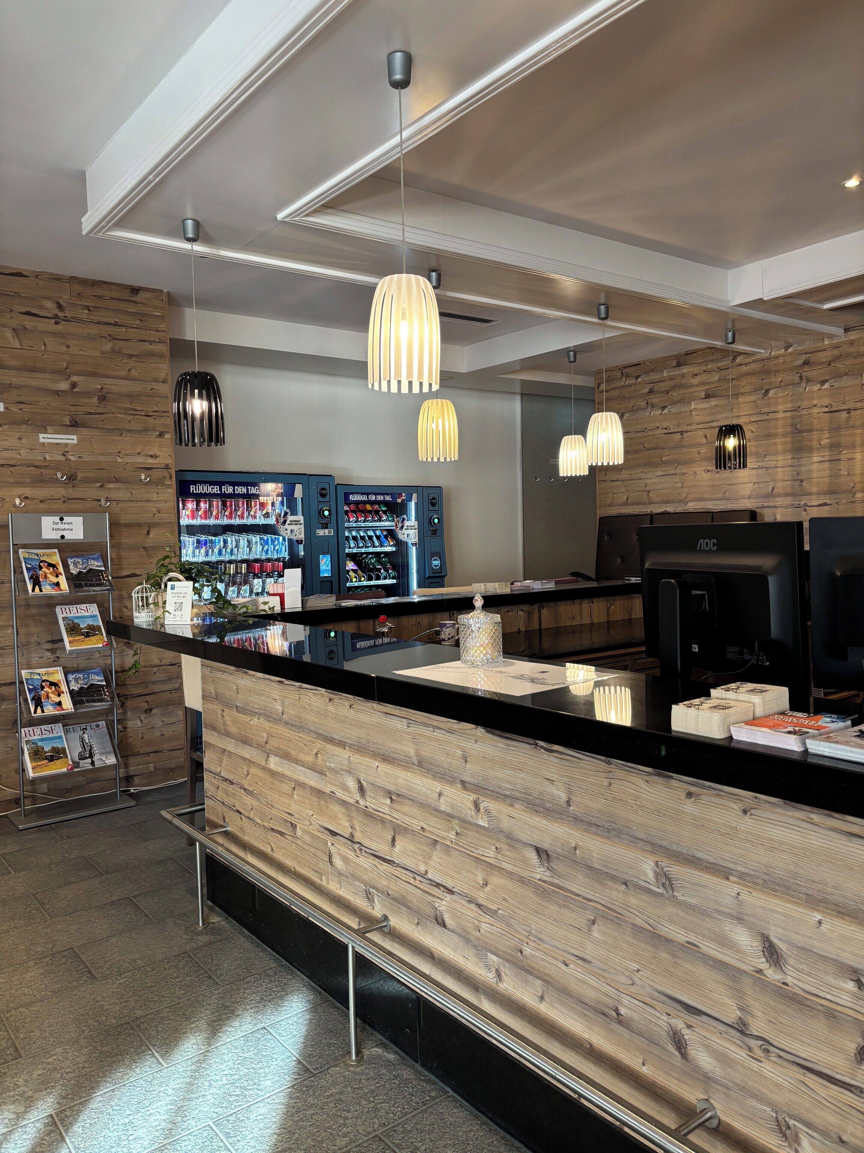 Un espace bar moderne agrémenté d’éléments en bois, d’un éclairage suspendu et d’un comptoir où sont exposées des boissons et des magazines.