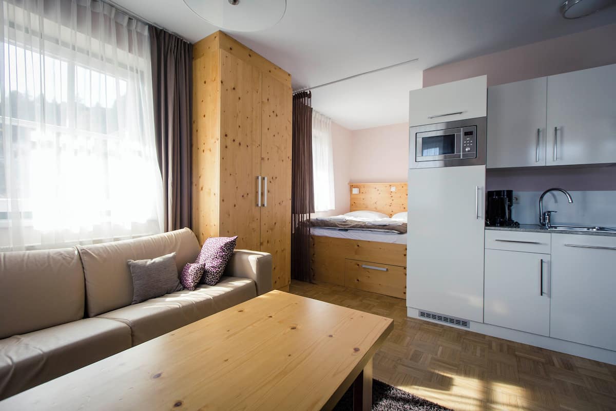 A compact living space with a sofa, a wooden table, a kitchenette with a microwave, and a bed with a wooden headboard.