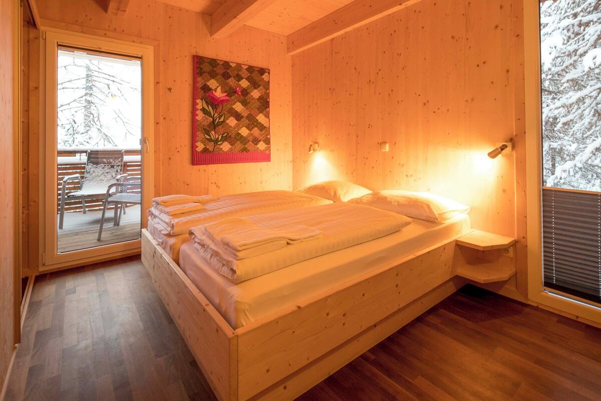 A wooden cabin bedroom with a bed, a quilted wall hanging, and a view of a snowy landscape through a window.
