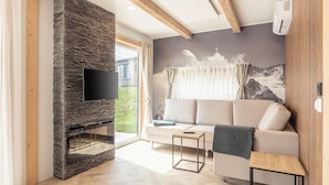 A modern living room with a stone fireplace, a large flat-screen TV, a sofa, and a window with a mountain view. - Chalets & Glamping Nassfeld by ALPS RESORTS (Kotschach-Mauthen)