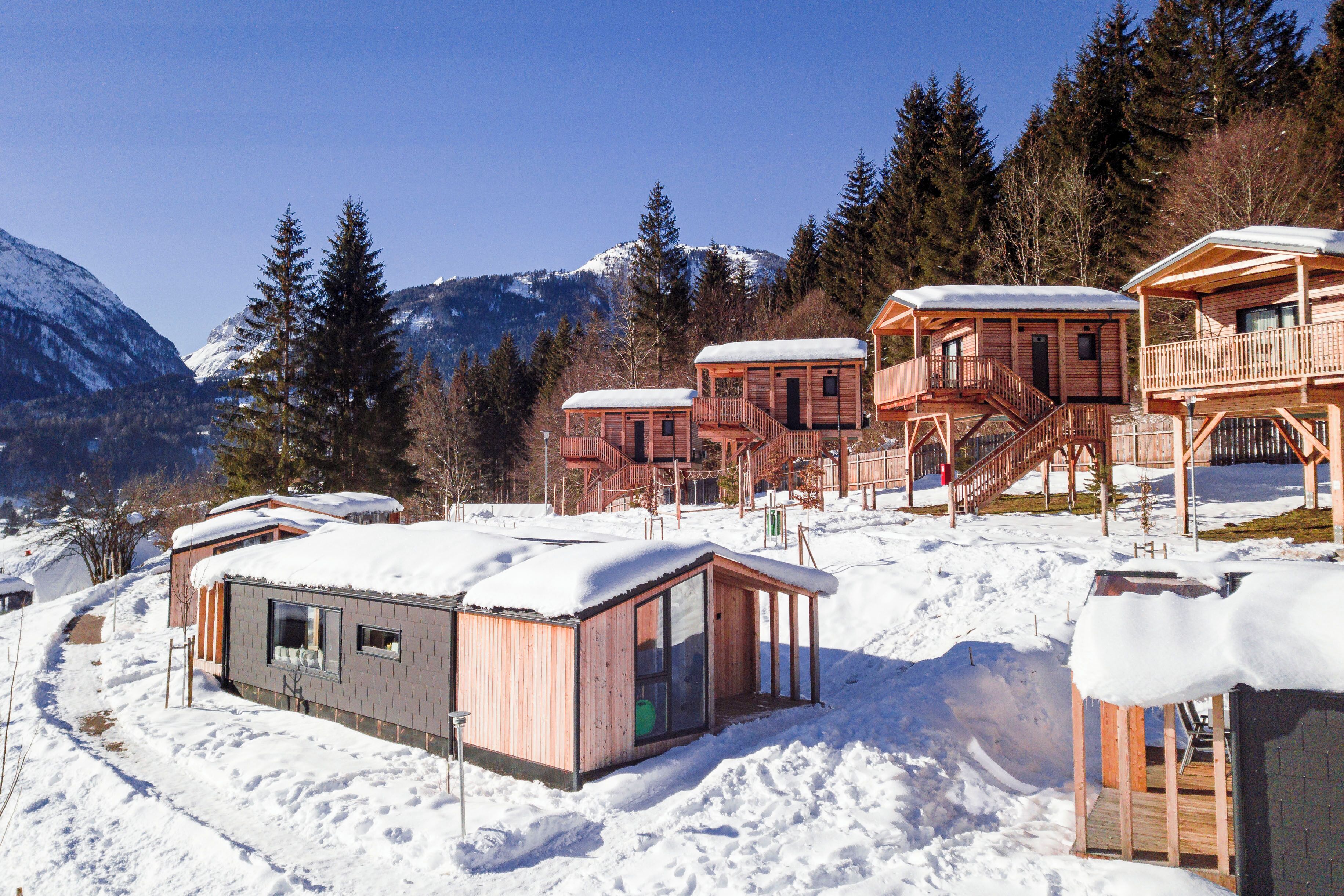 A cluster of wooden cabins with snow-covered roofs, set against a backdrop of snow-capped mountains and evergreen trees.