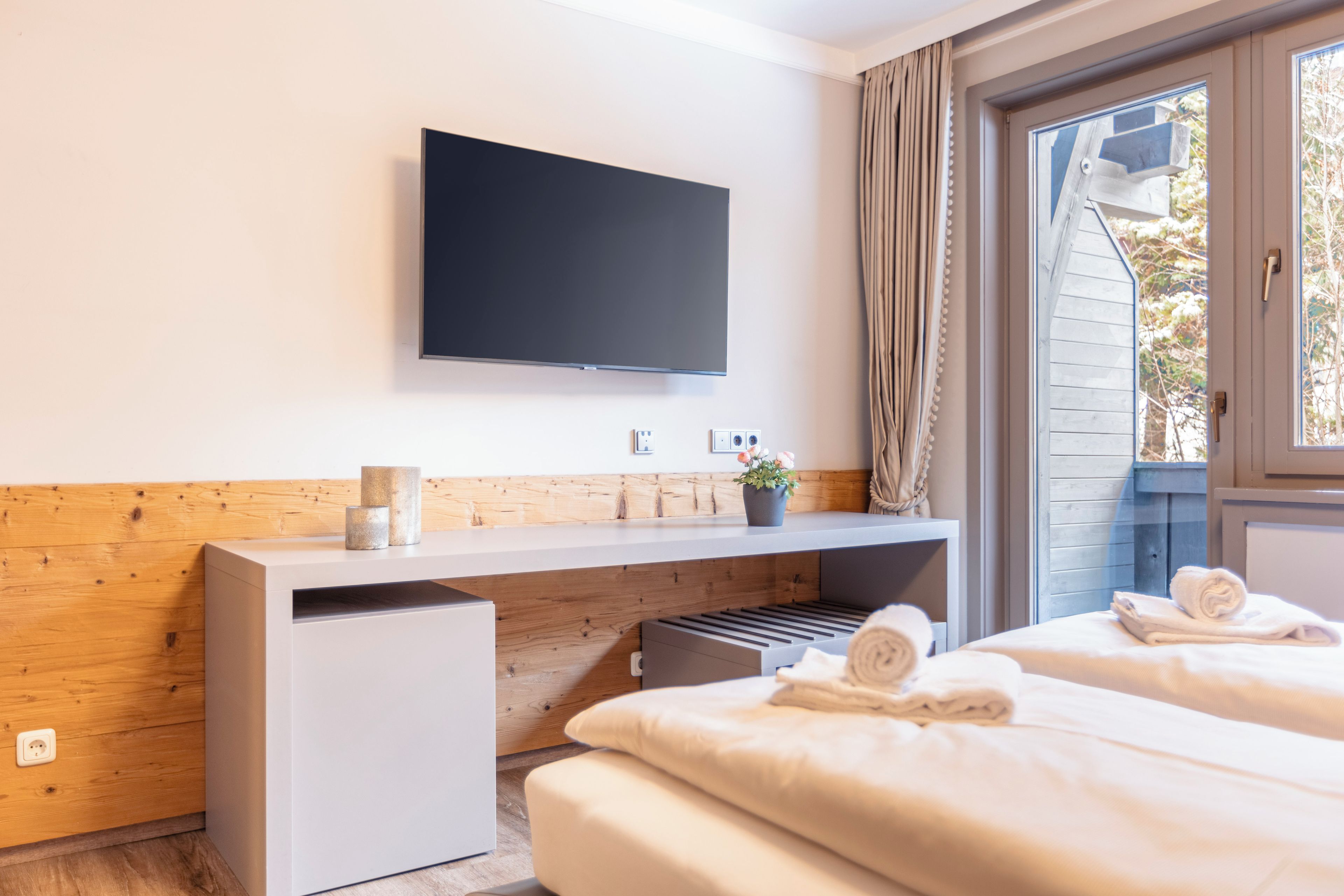 Een moderne hotelkamer met een flatscreen televisie aan de muur, een houten accentmuur, een klein bureau met een plant en twee bedden met handdoeken.