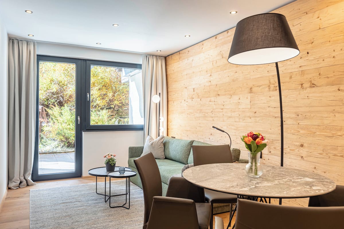 Ein modernes Zimmer mit einer Holzwand, einem runden Tisch mit Stühlen, einem Sofa, einer Lampe und Blick ins Grüne.