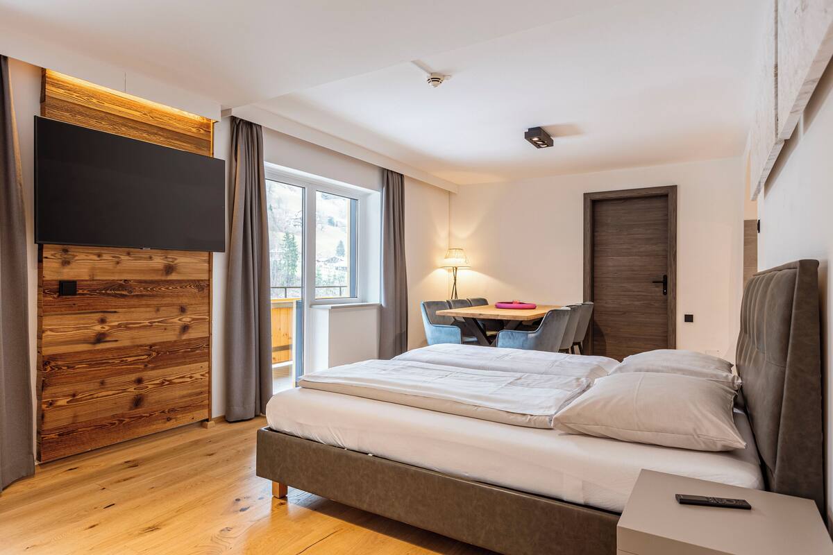 Ein modernes Schlafzimmer mit einem großen Bett, einem Schreibtisch mit Stuhl, einem Flachbildfernseher, der an einer Holzplatte befestigt ist, und einem Fenster mit Vorhängen.