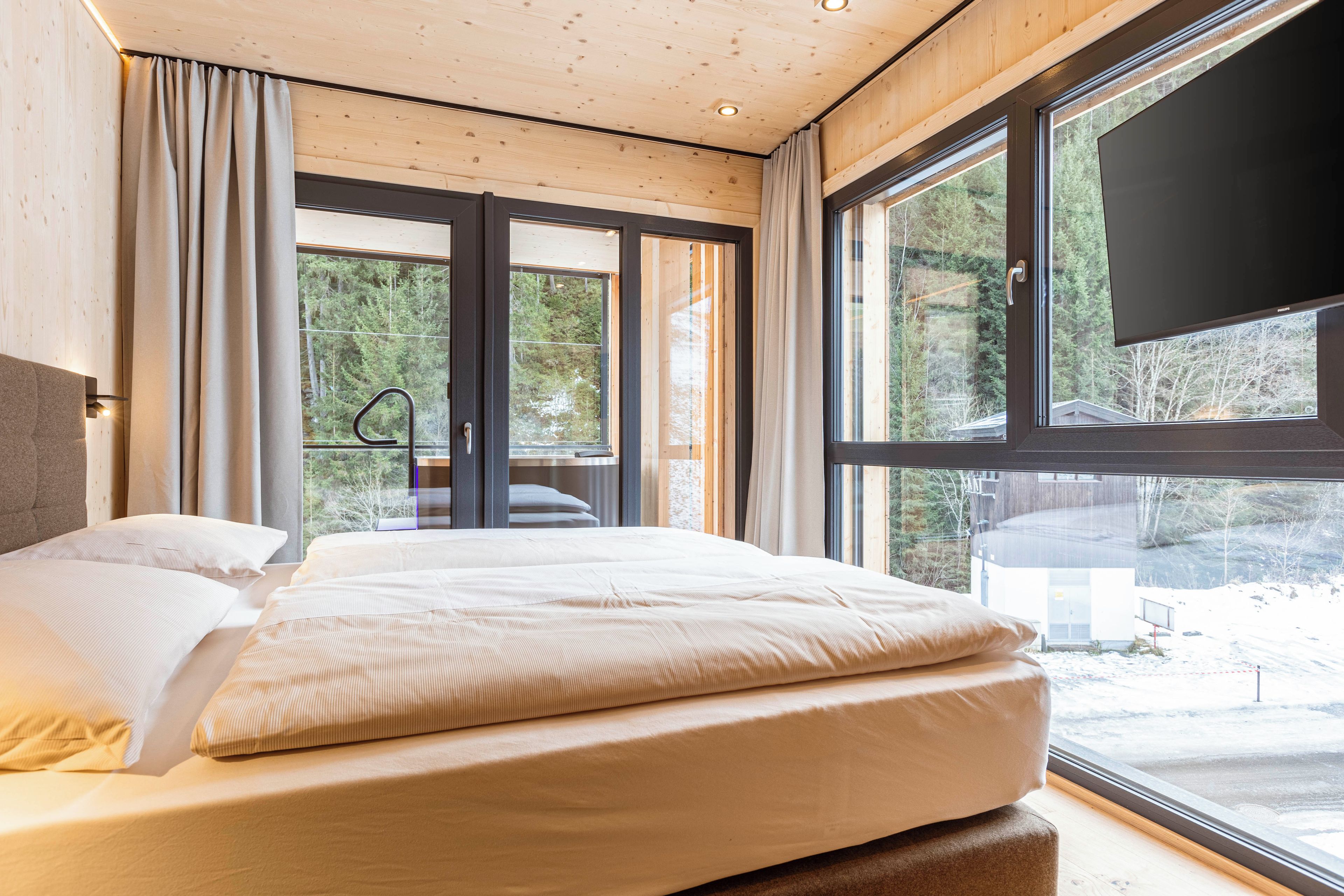 A modern bedroom with a large bed, a TV mounted on the wall, and a view of the outdoors.