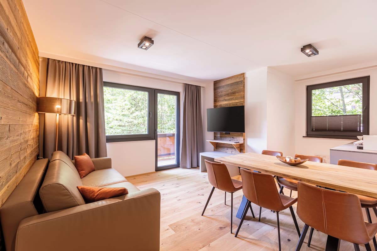 Ein modernes Wohnzimmer mit einer Holzvertäfelung, einer Couch, einem Esstisch mit Stühlen, einem Flachbildfernseher und großen Fenstern mit Vorhängen.