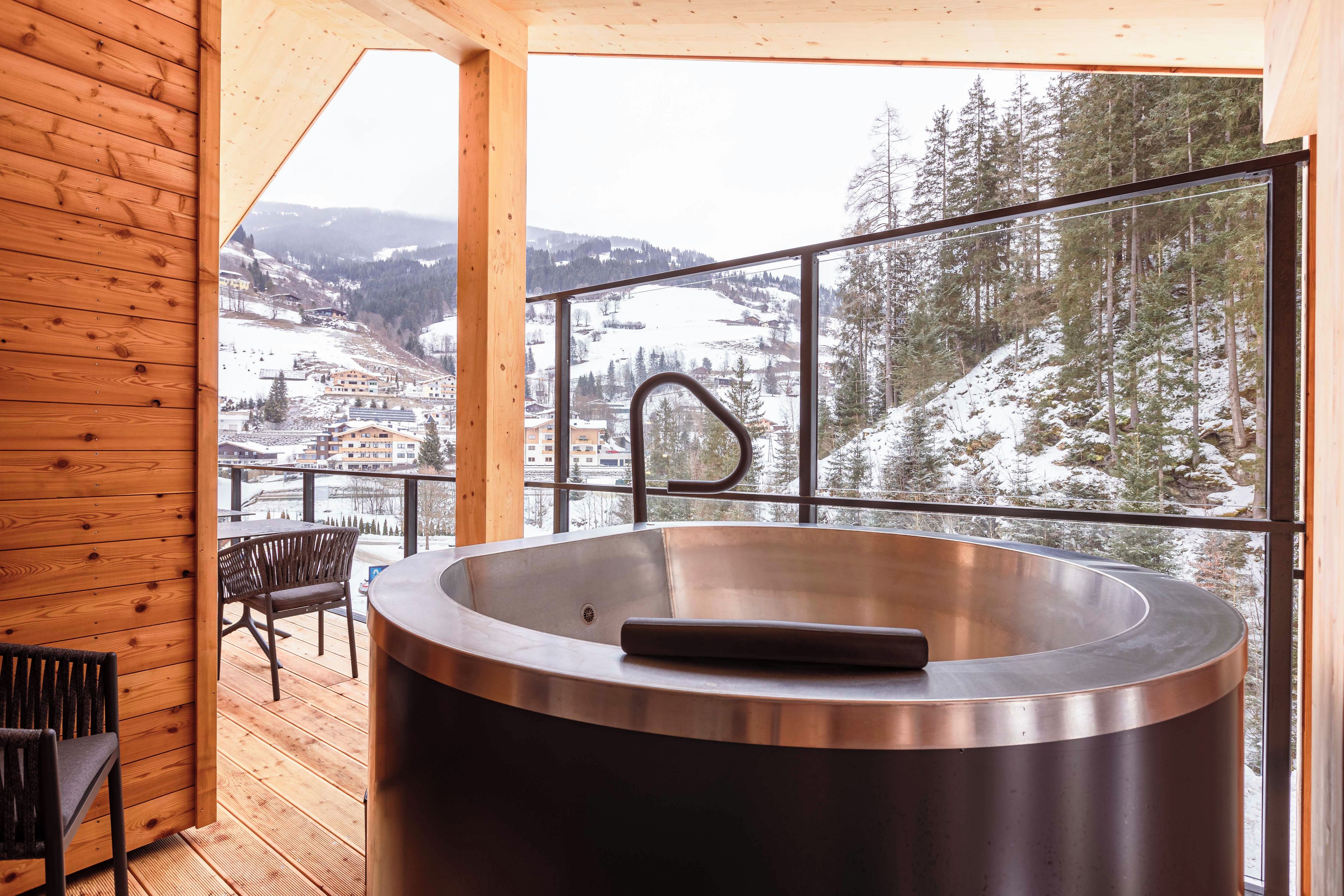 A modern bathroom with a large bathtub, wooden deck, and a scenic view of a snowy mountain town.