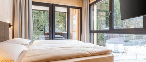 A modern bedroom with a large bed, a TV mounted on the wall, and a view of the outdoors.