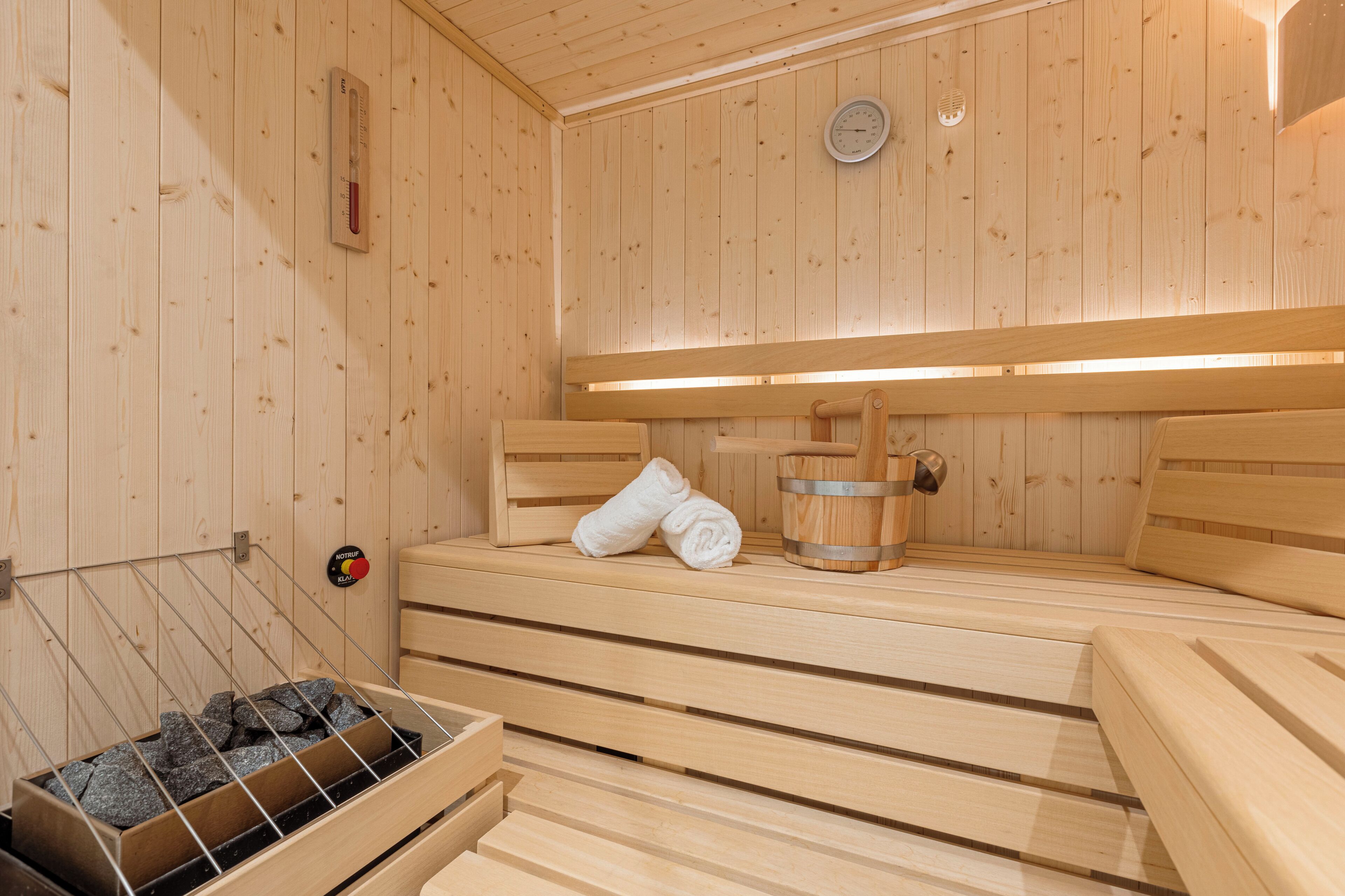 A wooden sauna with benches, a thermometer, a clock, and a bucket.