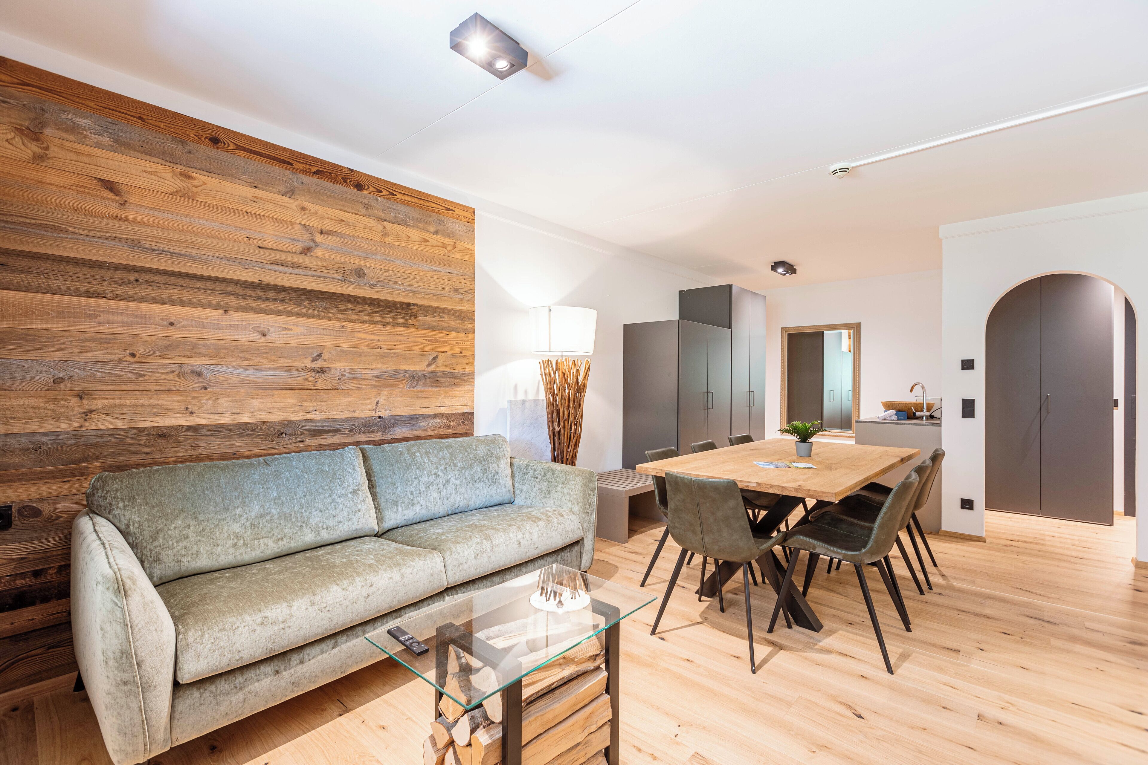 A modern living room with a wooden accent wall, a glass coffee table, a sofa, and a dining area with a wooden table and chairs.