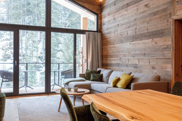 A cozy room with a large window, a wooden wall, a sofa, a table, and a chair. - Saalbach Suites by ALPS RESORTS (Saalbach-Hinterglemm)
