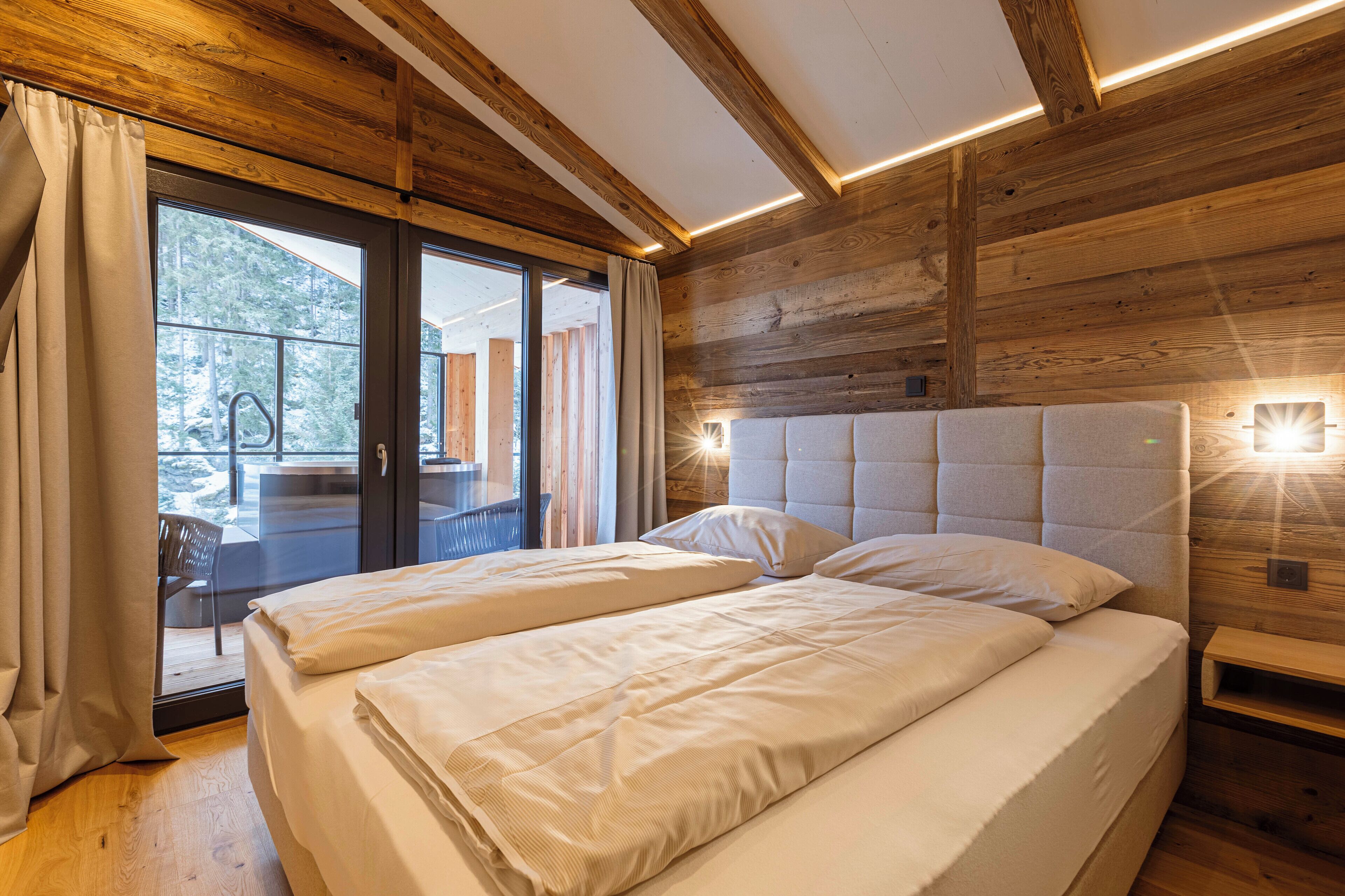 Una cama doble con cabecera de madera y una mesita de noche, una puerta corrediza de vidrio que da a un espacio exterior, y un techo de madera con vigas a la vista.