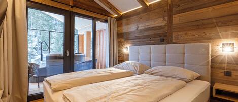 Una cama doble con cabecera de madera y una mesita de noche, una puerta corrediza de vidrio que da a un espacio exterior, y un techo de madera con vigas a la vista.