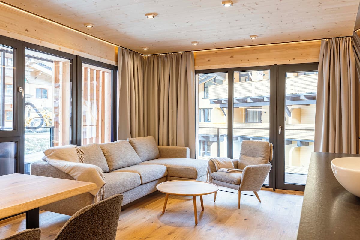 Ein modernes Wohnzimmer mit einer Couch, Sesseln, einem runden Tisch und großen Fenstern mit Vorhängen.
