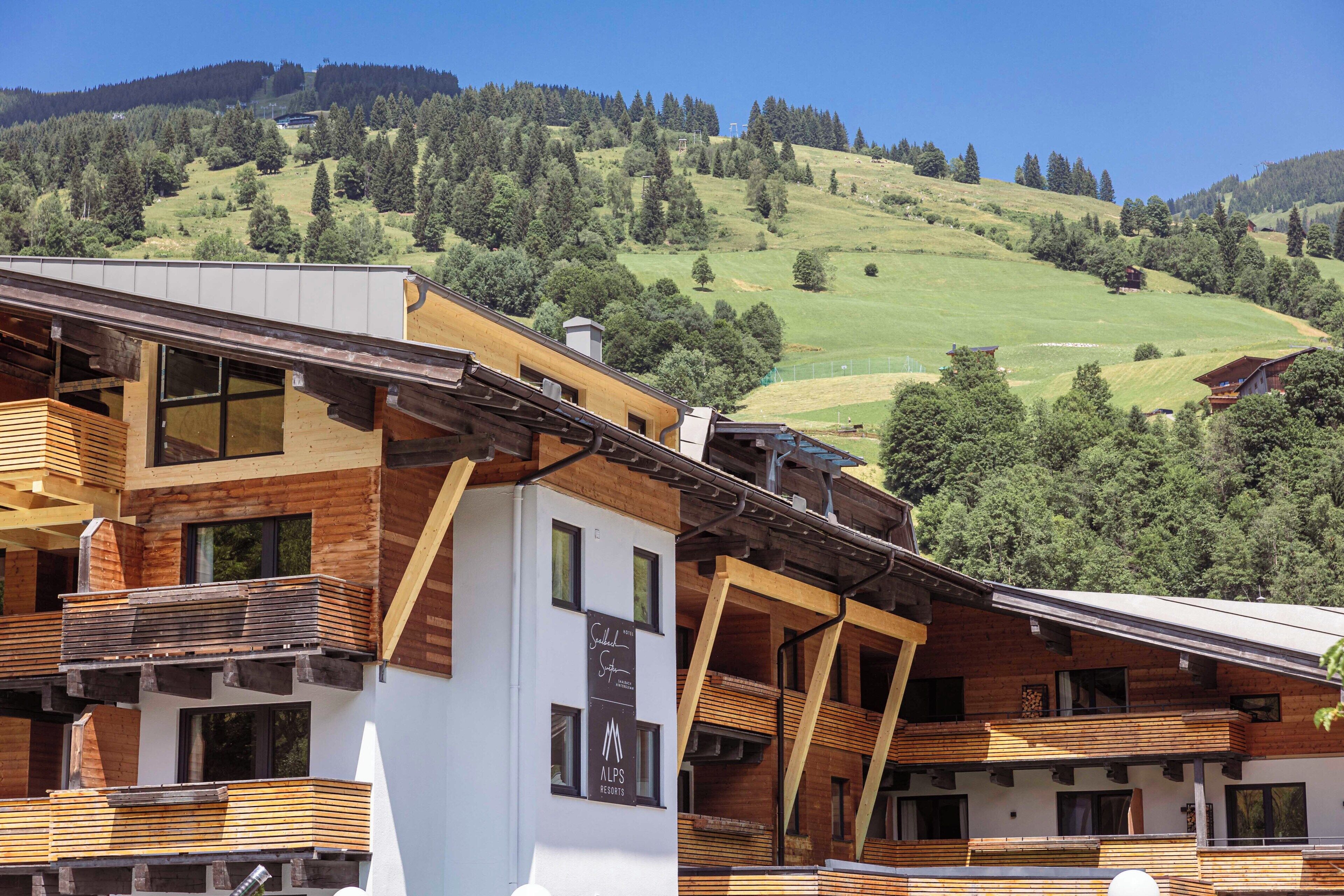 A modern building with wooden balconies, a sign for 'Alps Resorts,' and a view of a green hillside.