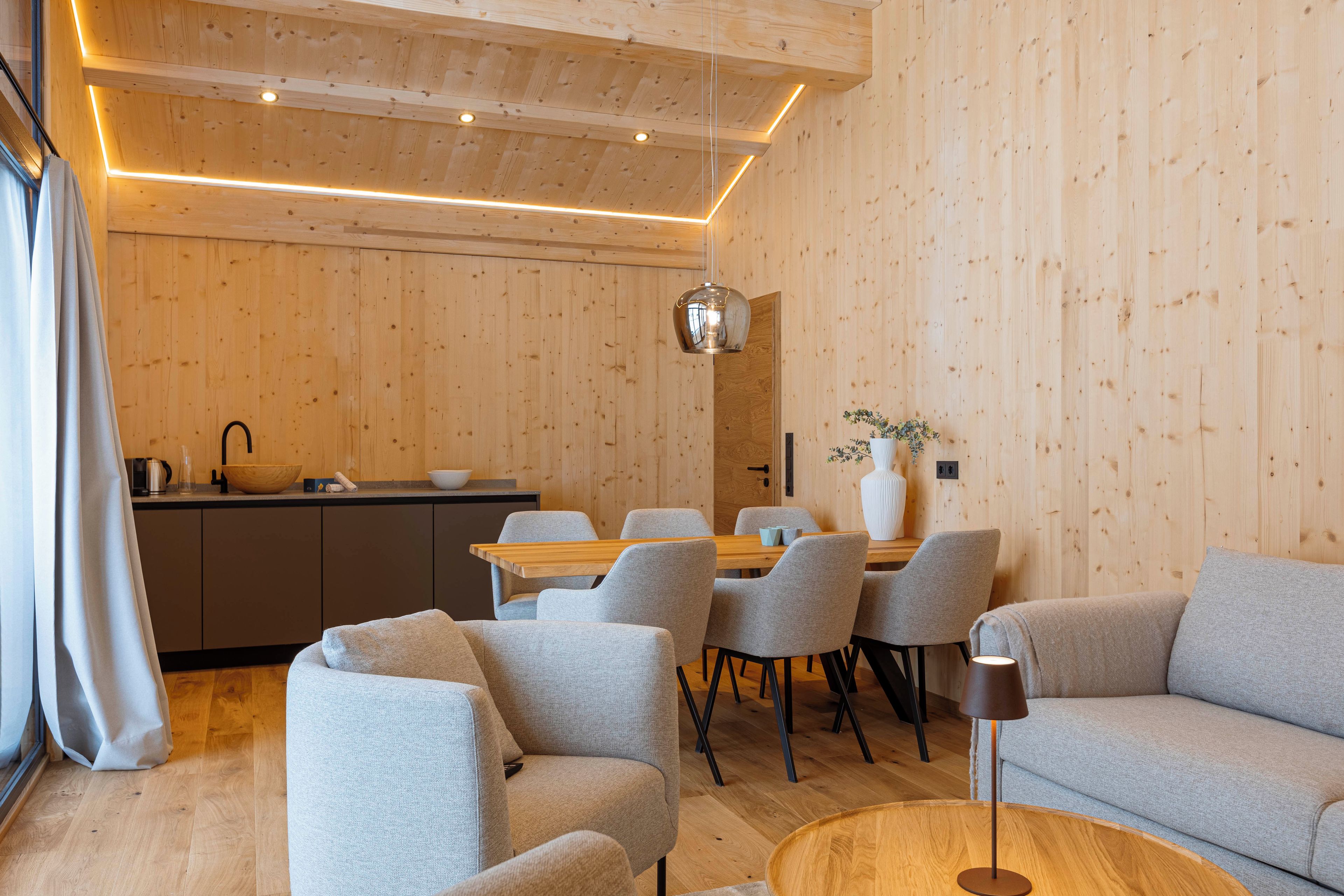 A modern interior with a wooden ceiling, a dining area with grey chairs, a sofa, and a round wooden table.