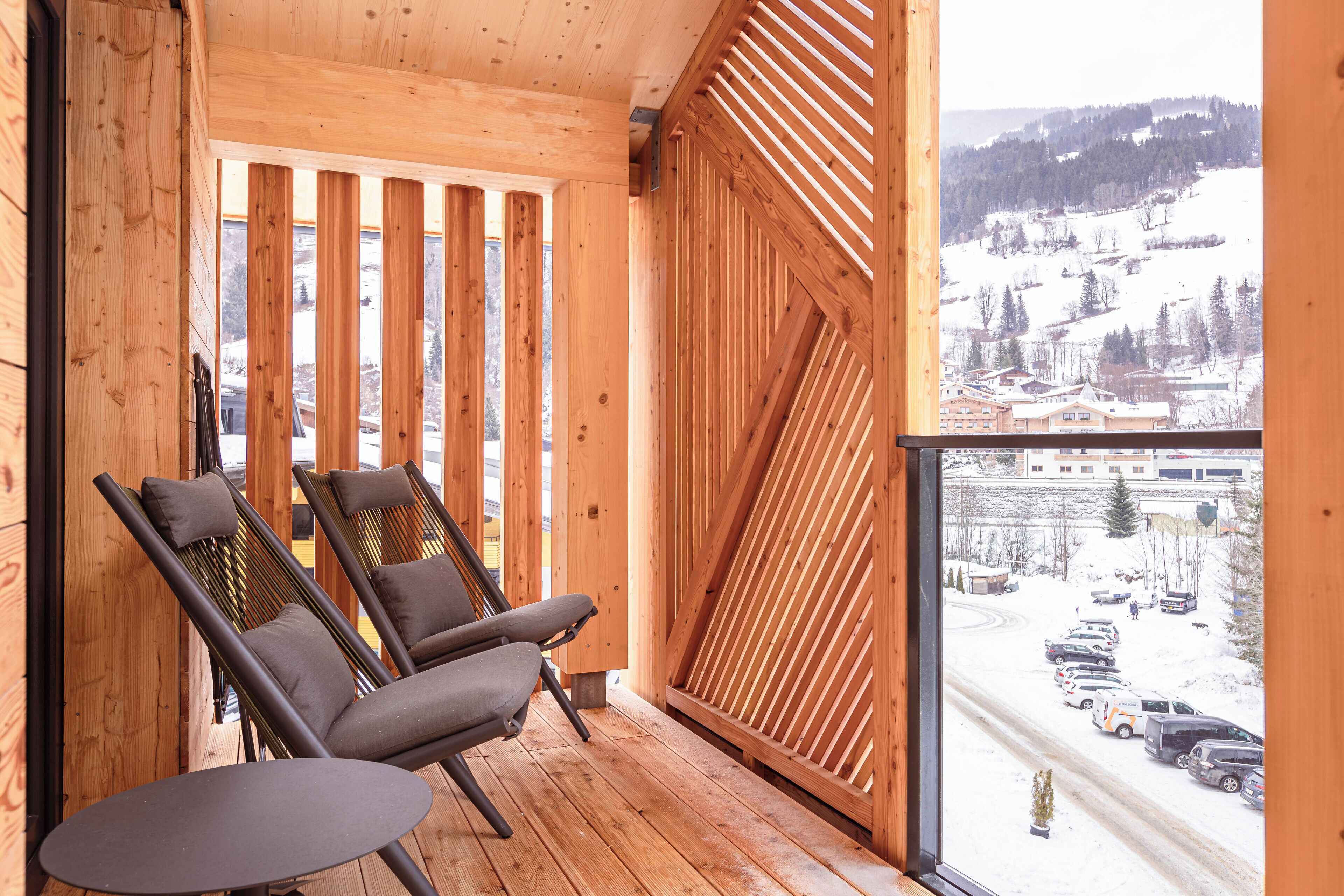 A wooden balcony with two chairs and a table, overlooking a snowy landscape with buildings and parked cars.