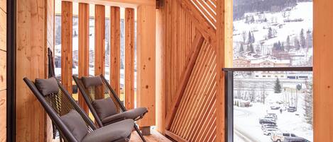 A wooden balcony with two chairs and a table, overlooking a snowy landscape with buildings and parked cars.