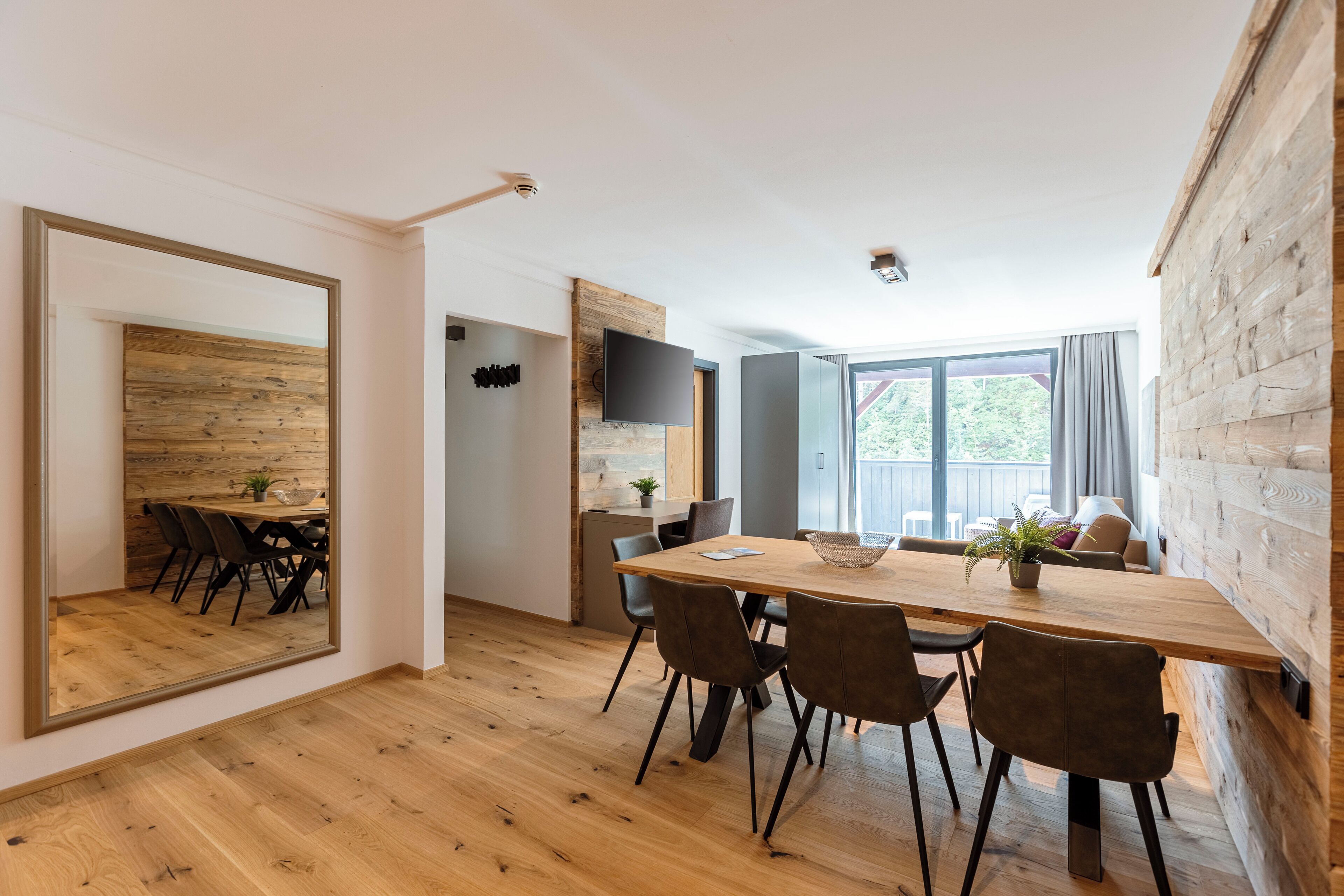 A modern dining area with a wooden table and chairs, a large mirror, and a television on the wall.