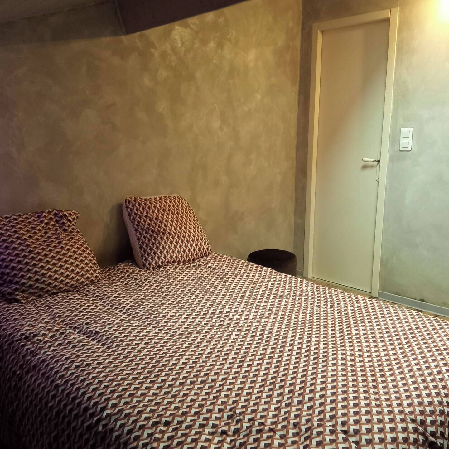 Een slaapkamer met een bed met een patroon, twee kussens, een nachtkastje en een deur met lichtschakelaar.