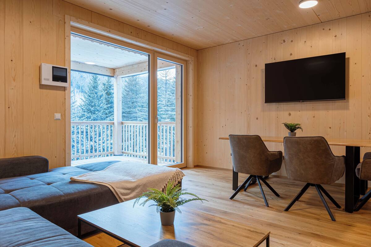 Une pièce moderne avec un intérieur en bois, un canapé, une table à manger avec des chaises, un téléviseur fixé au mur et une grande fenêtre donnant sur des arbres couverts de neige.