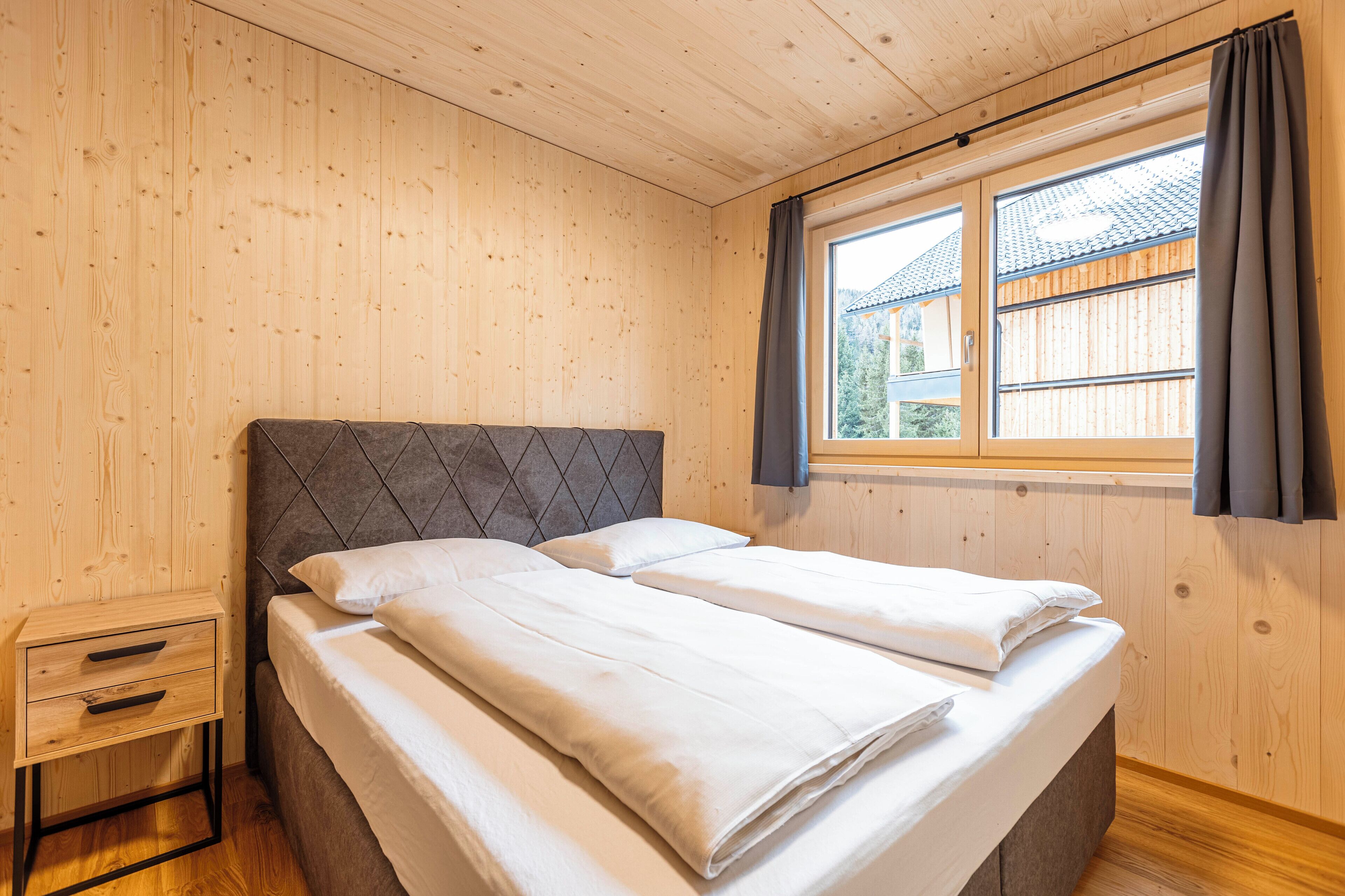 A bedroom with a wooden headboard, a small bedside table, and a window with curtains.